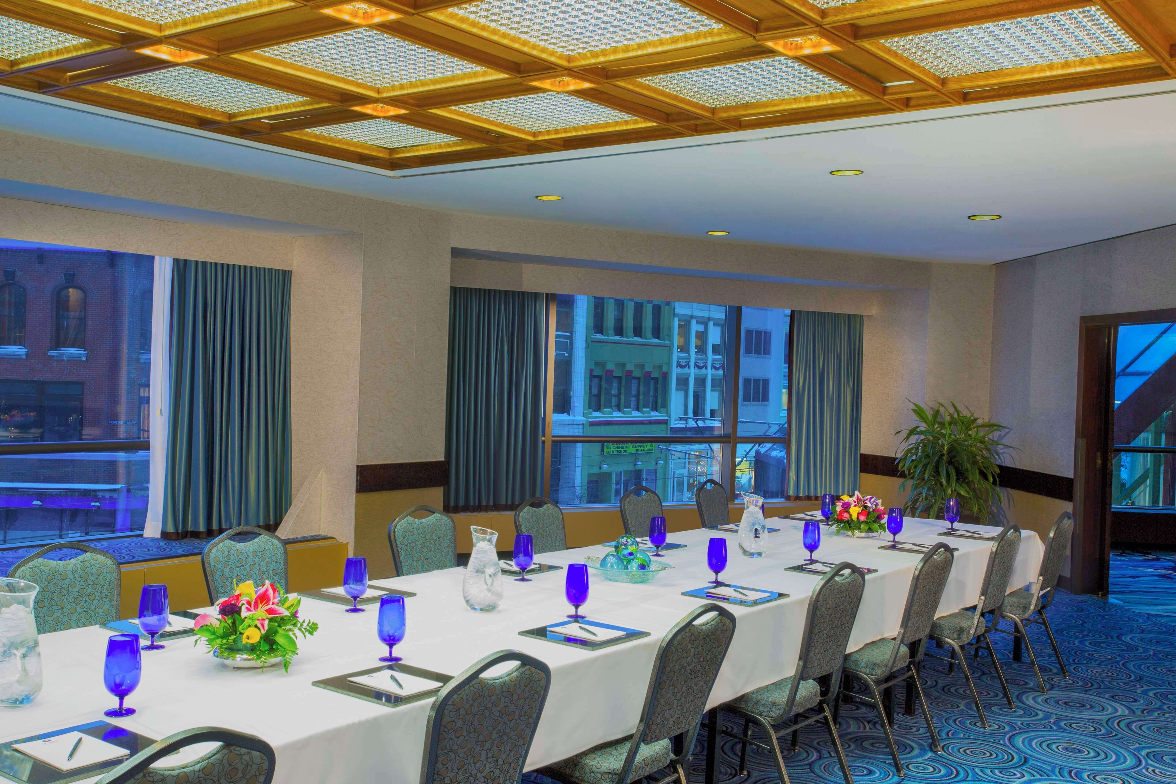 Un salón de conferencias con una larga mesa dispuesta para una reunión, ventanas amplias con cortinas y vista a un edificio exterior.