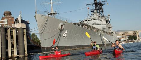 A large naval ship docked at a pier with three kayakers in red kayaks.