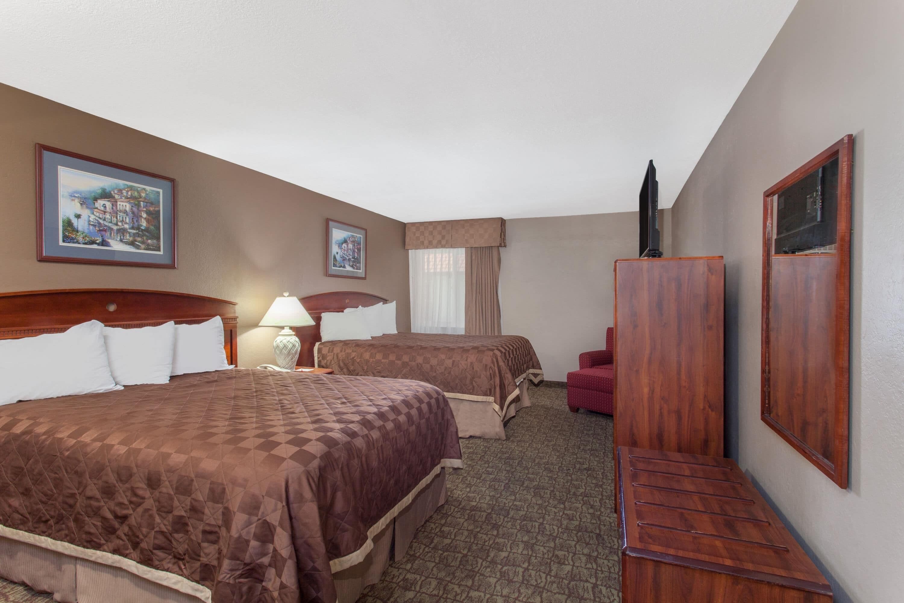 A hotel room with two beds, a desk, a chair, a lamp, and two framed pictures on the wall.