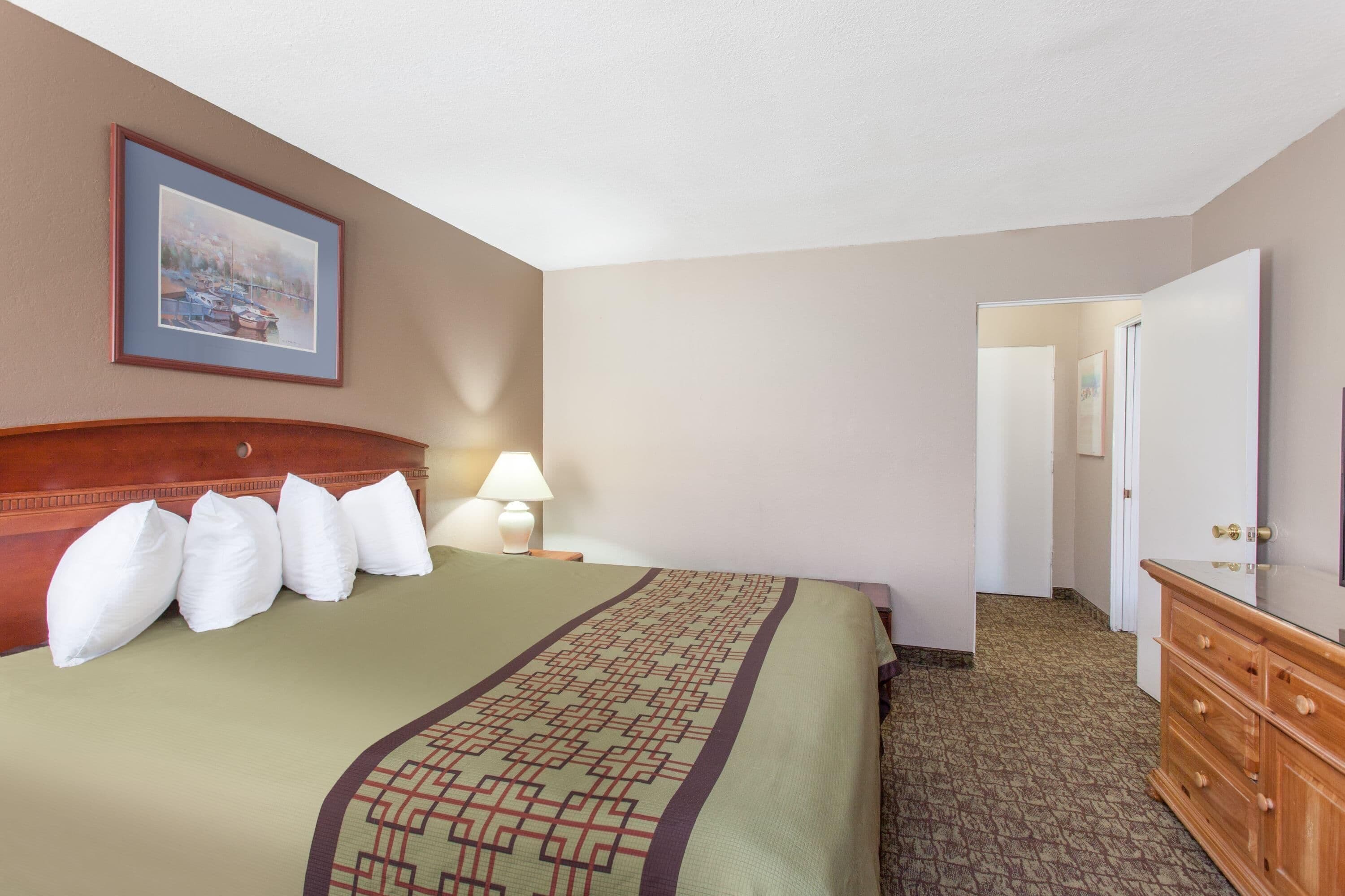 A hotel room with a bed, pillows, a lamp, a dresser, a mirror, and a painting on the wall.