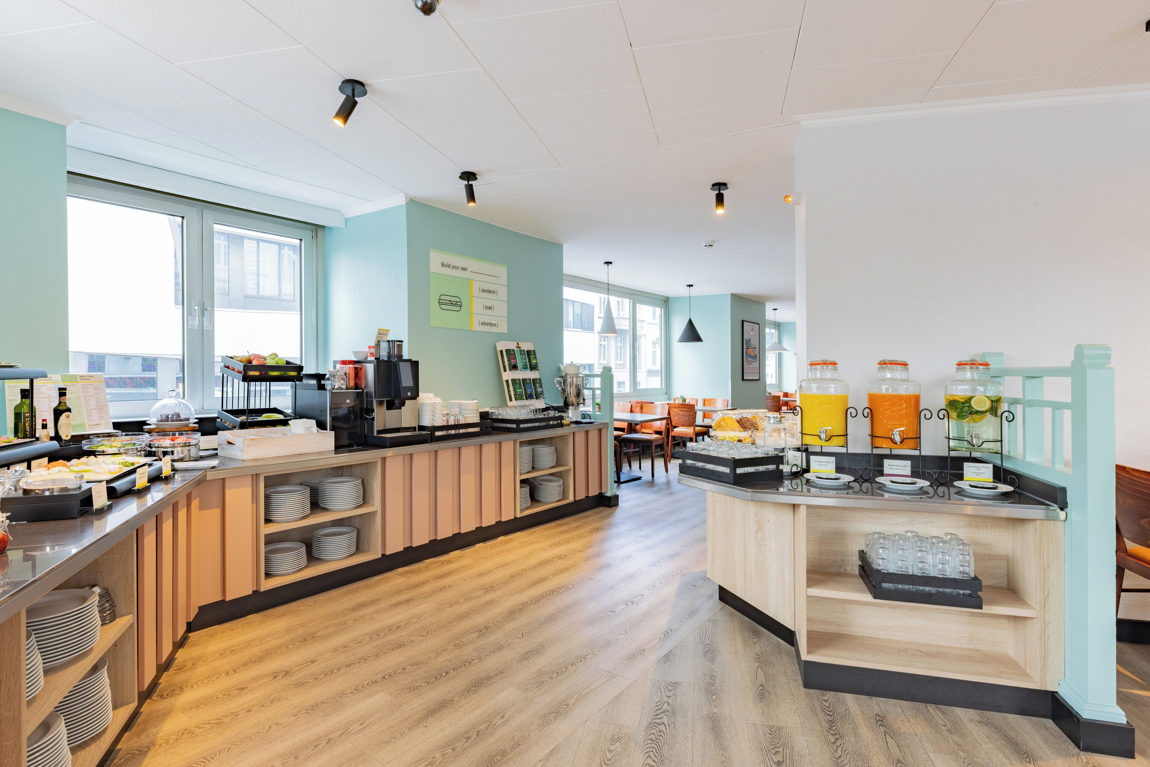 A modern breakfast bar with a variety of food and drink options, wooden flooring, and large windows providing natural light.