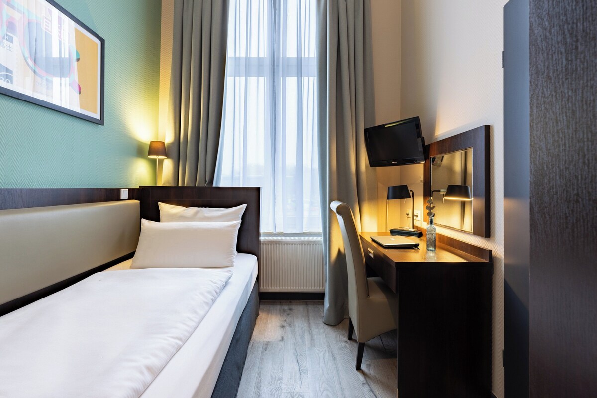 Habitación de hotel con cama, escritorio, silla, televisor y ventana con cortinas.