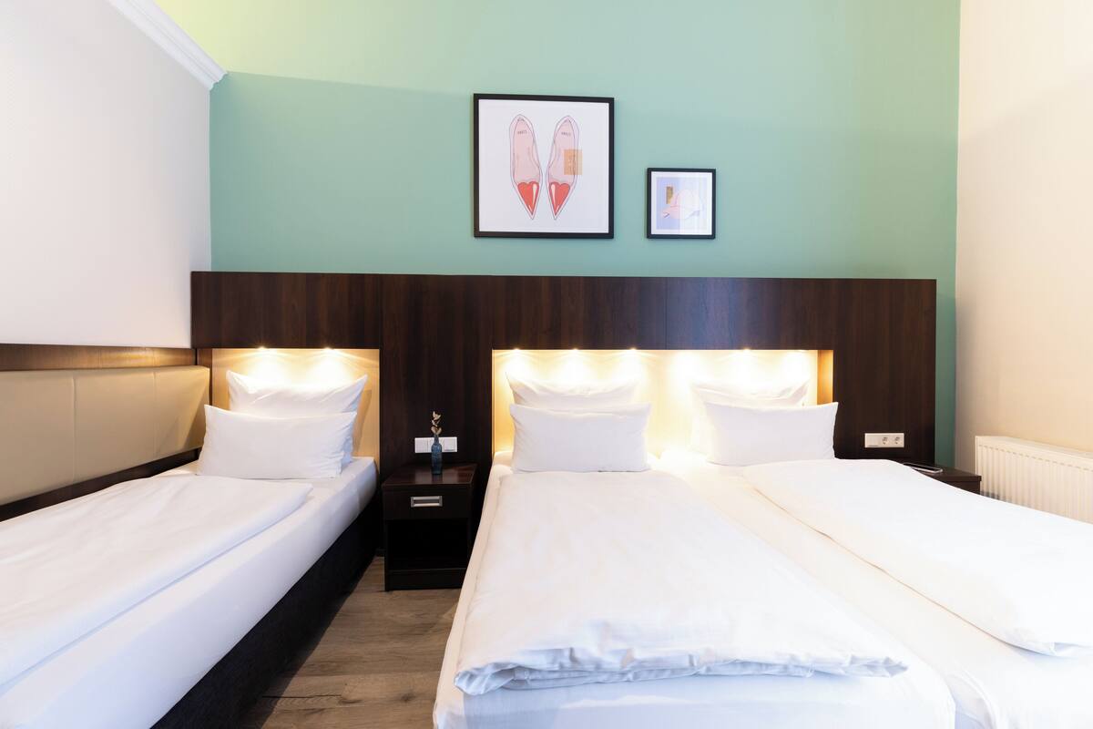 A hotel room with two single beds, a wooden headboard, a nightstand with a lamp, and artwork on the wall.