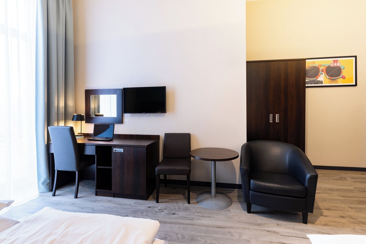 Habitación de hotel con cama, escritorio con computadora, silla, mesita y televisor en la pared.