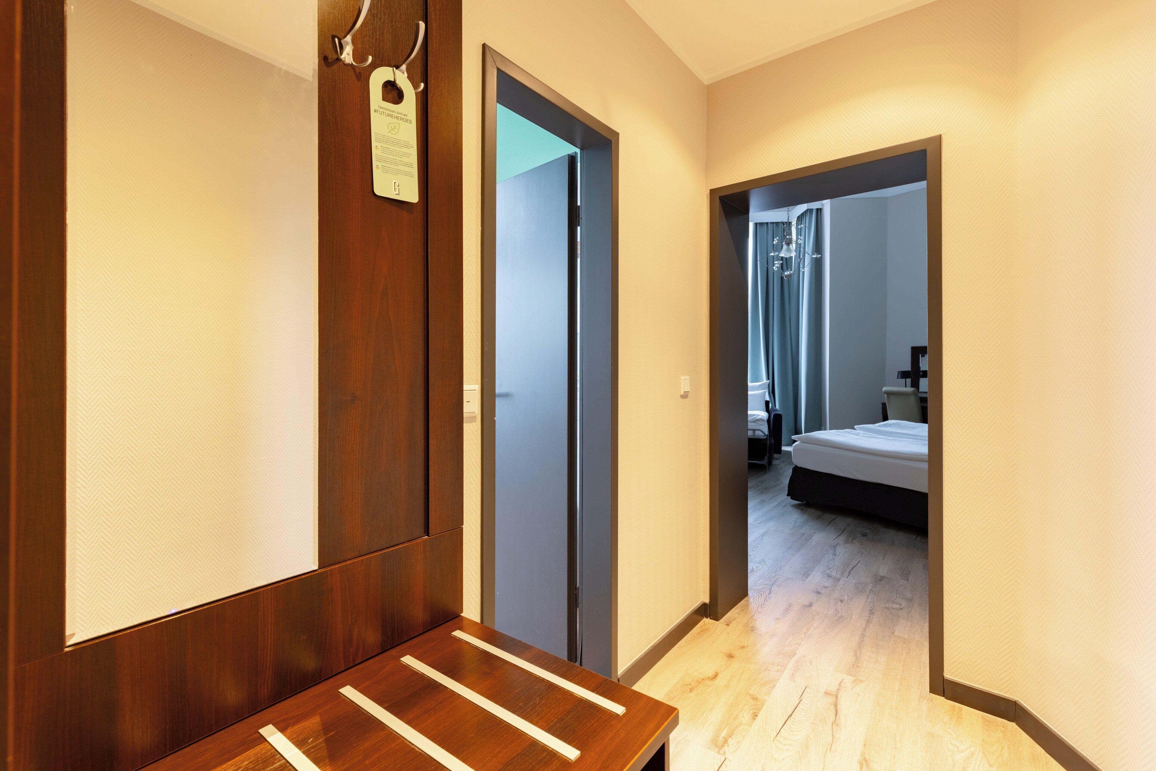 A hotel hallway with a wooden bench, a door with a keycard holder, and a glimpse of a bedroom with a bed and a nightstand.