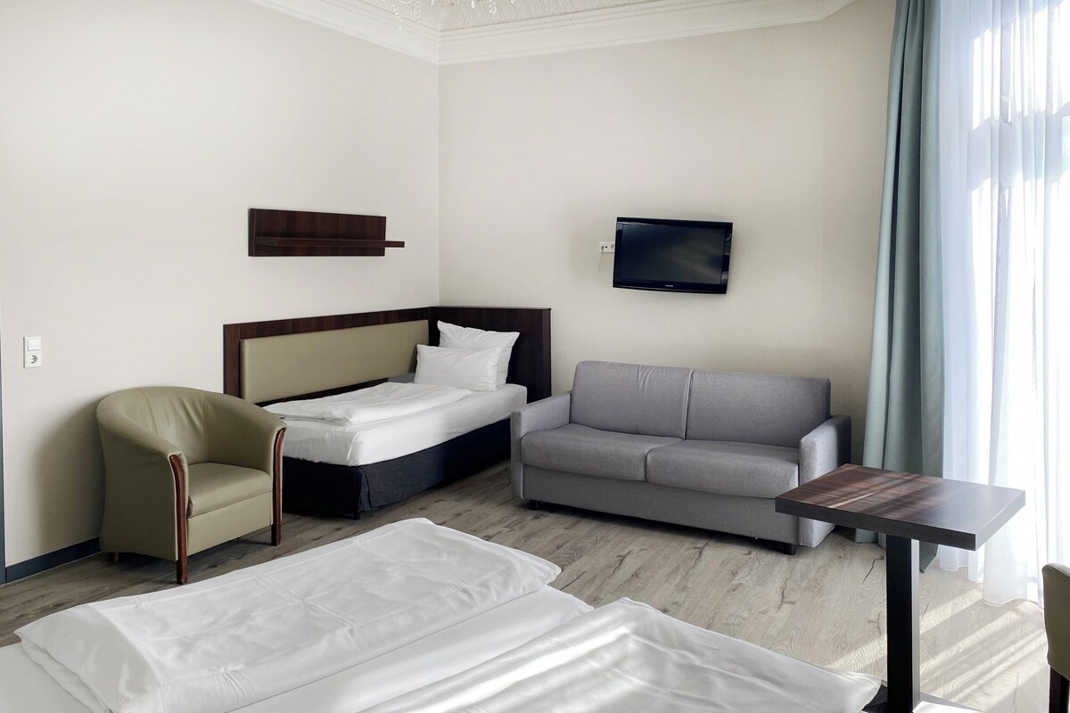 Una habitación de hotel con cama, sofá, sillón, televisor y una mesita.