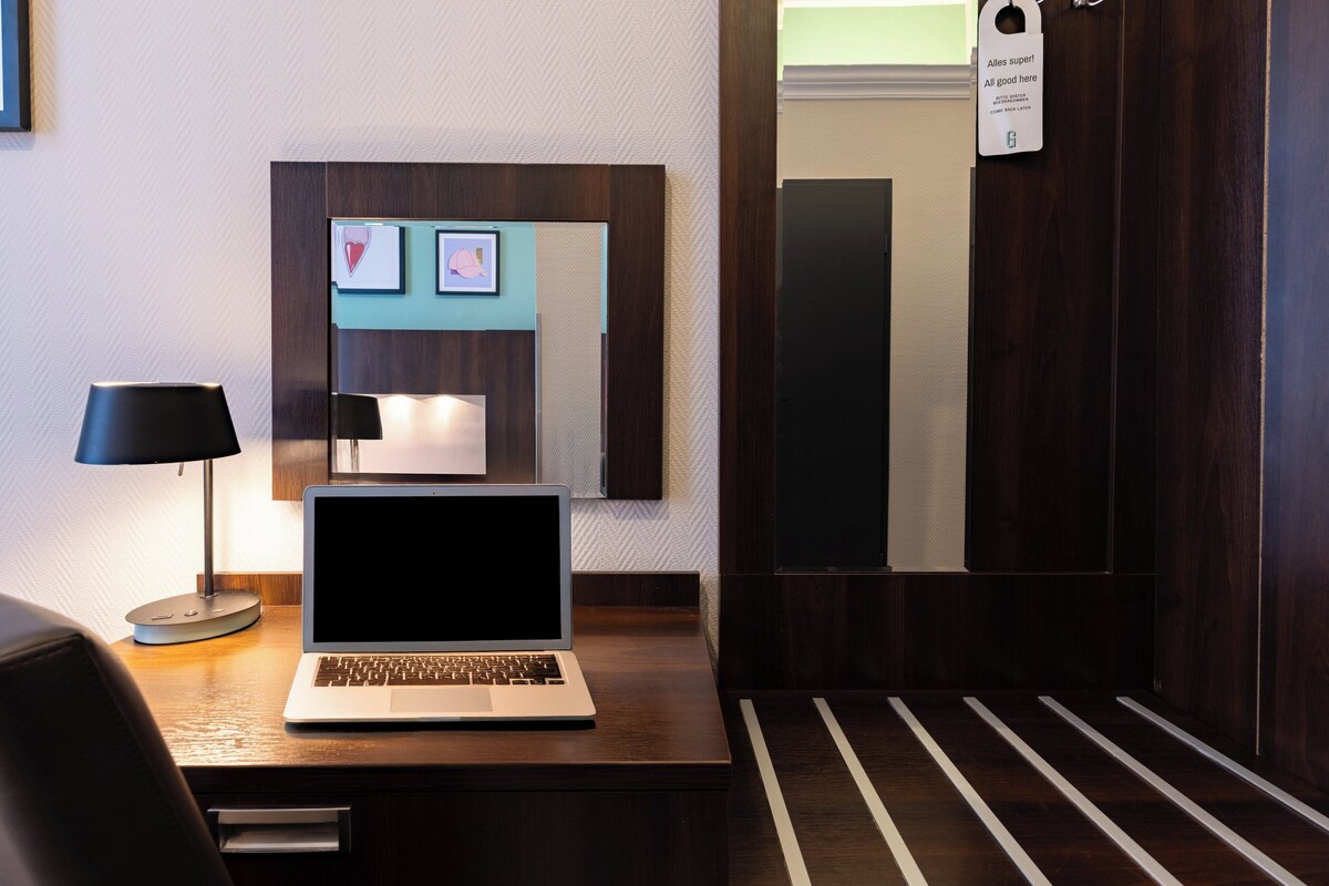 Habitación de hotel con escritorio de madera, una computadora portátil, lámpara y un cuadro en la pared.
