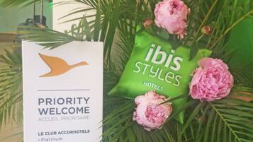 A 'Priority Welcome' sign with a bird logo, a green 'ibis Styles Hotels' bag, and pink flowers.
