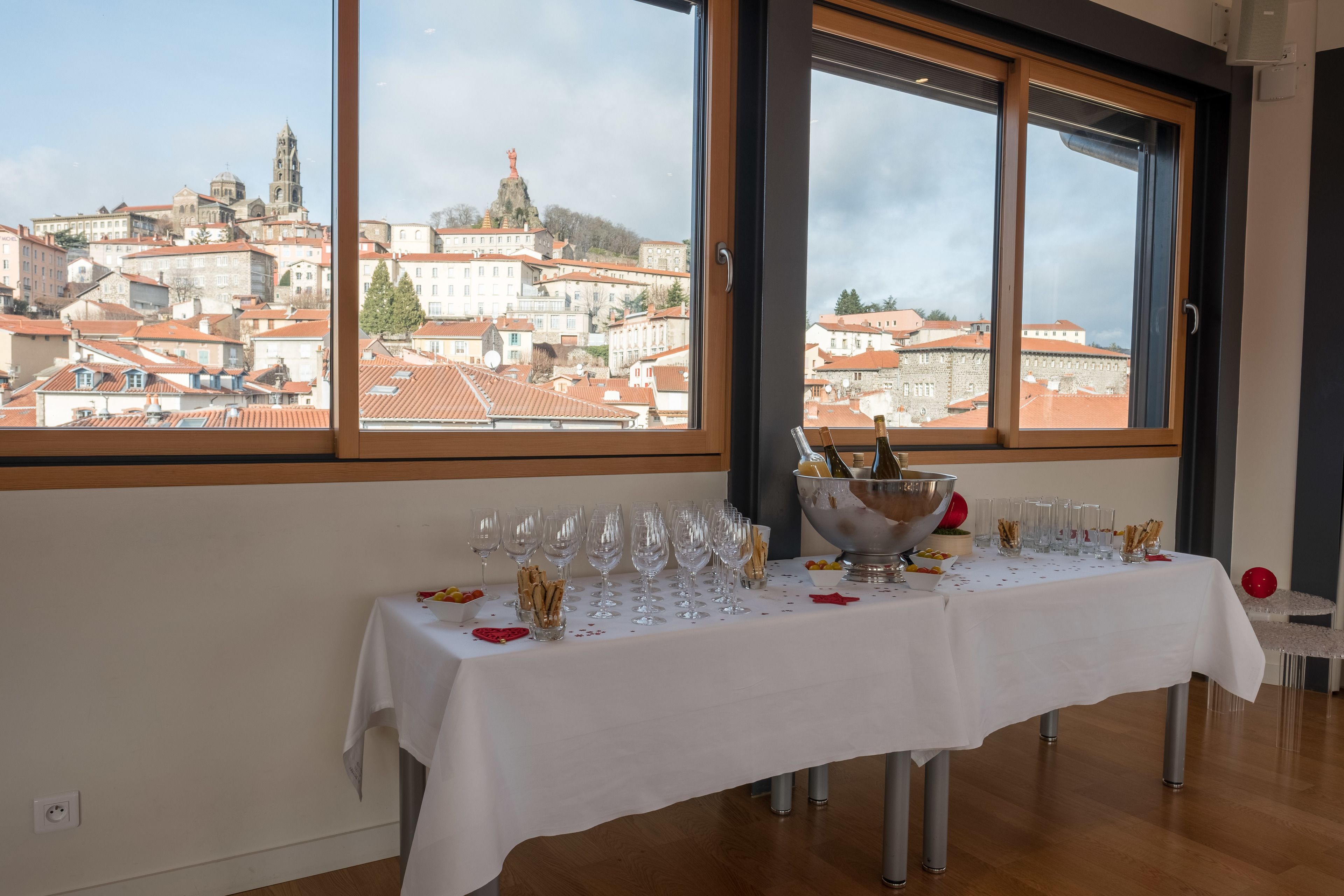 A table set with wine glasses and a bottle, a bowl of snacks, and a red balloon, with a city view through large windows.