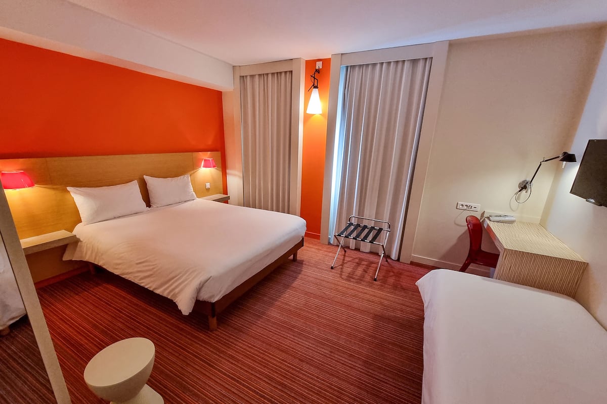 Une chambre d’hôtel avec un grand lit, des lampes de chevet, un bureau avec une chaise, une petite table et une fenêtre avec des rideaux.