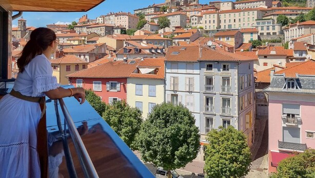 Un balcon donnant sur une ville historique, avec son architecture traditionnelle, un clocher d’église imposant et une statue sur une colline.