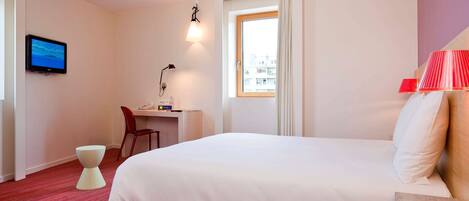 Habitación de hotel con cama, escritorio y silla, televisor, lámpara y ventana con cortinas.