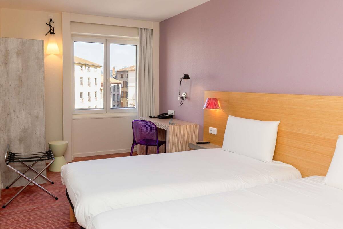 Une chambre d’hôtel avec un lit, un bureau, une chaise, une lampe et une vue sur les immeubles par la fenêtre.