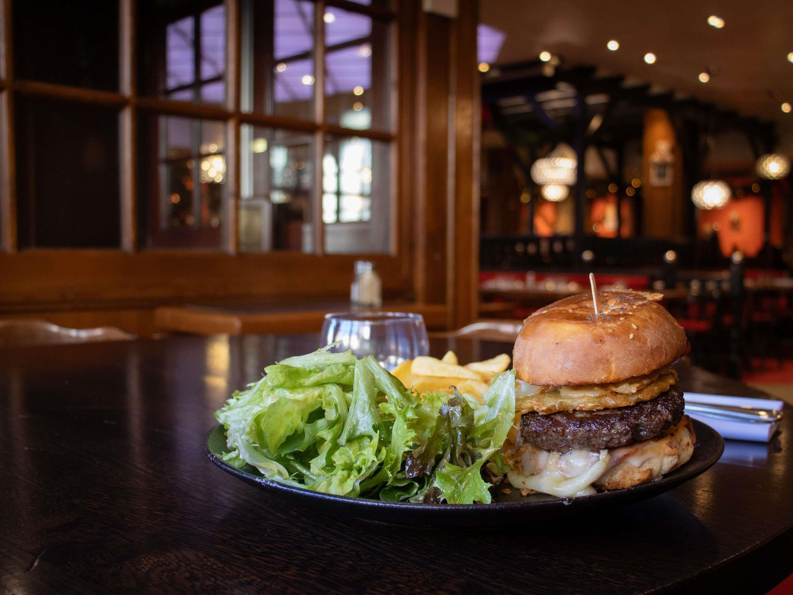 Ein Burger mit Pommes und Salat auf einem Teller, platziert auf einem Tisch in einem Restaurant mit Holzvertäfelungen und großen Fenstern.