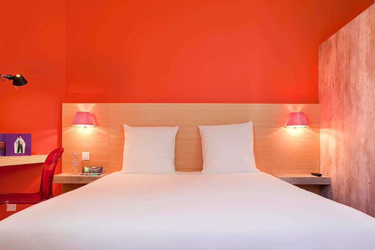 A hotel room with a large bed, two bedside lamps, a wooden headboard, a red chair, and a small table with a bottle and a book.