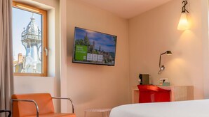Une chambre d’hôtel comprenant un lit, une chaise, un bureau, une télévision et une fenêtre donnant sur un immeuble.