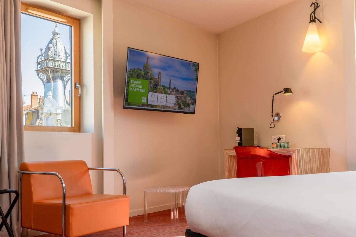 Une chambre d’hôtel comprenant un lit, une chaise, un bureau, une télévision et une fenêtre donnant sur un immeuble.