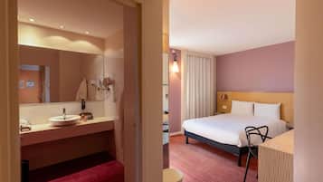 Habitación de hotel con cama, mesita de noche, espejo, lavabo y un banco rojo.