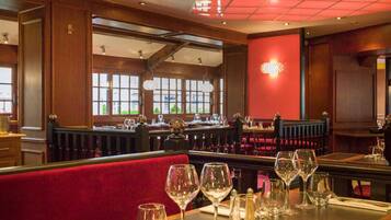 Ein Restaurant Innenraum mit Holzwänden, rot gepolsterten Sitzecken und einer roten Akzentwand. Die Tische sind mit Weingläsern und Besteck gedeckt.