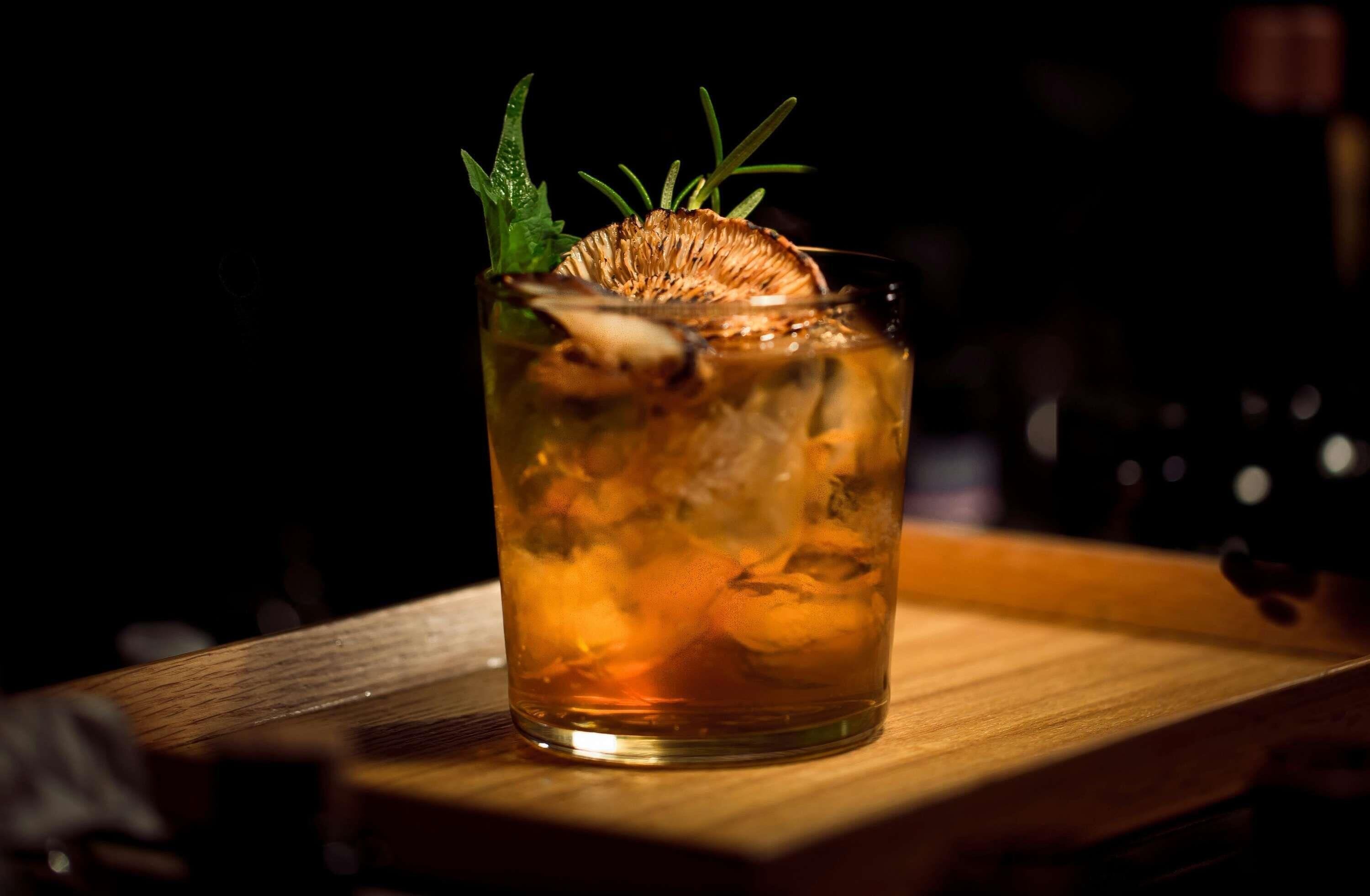 A glass of amber-colored beverage with ice cubes and a sprig of rosemary on a wooden bar counter.