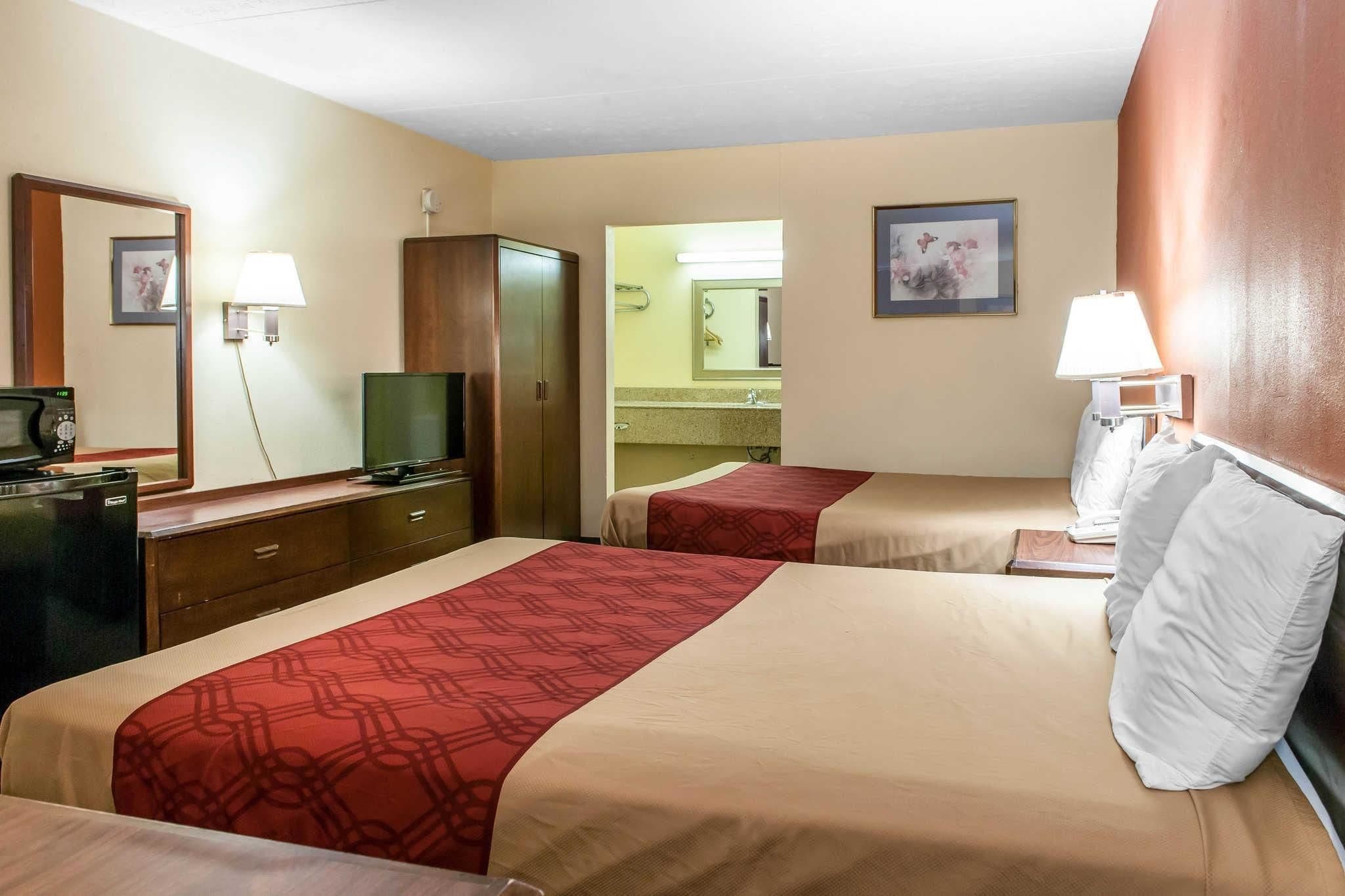 Habitación de hotel con dos camas, un escritorio con televisor, un espejo y un baño visible a través de una puerta abierta.