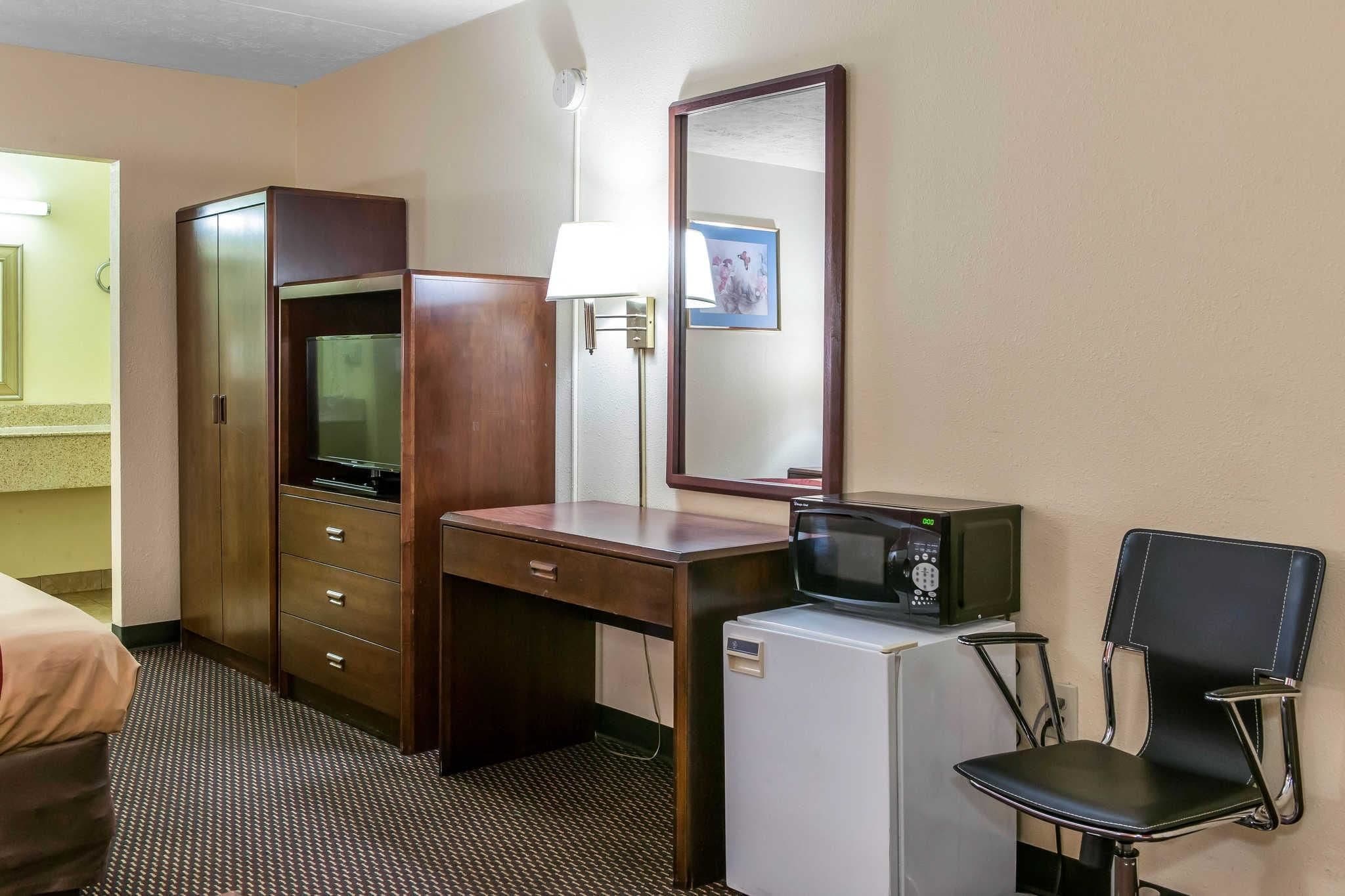 A hotel room with a wooden desk, a television, a microwave, a refrigerator, a chair, a lamp, and a mirror.
