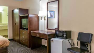 A hotel room with a wooden desk, a television, a microwave, a refrigerator, a chair, a lamp, and a mirror.