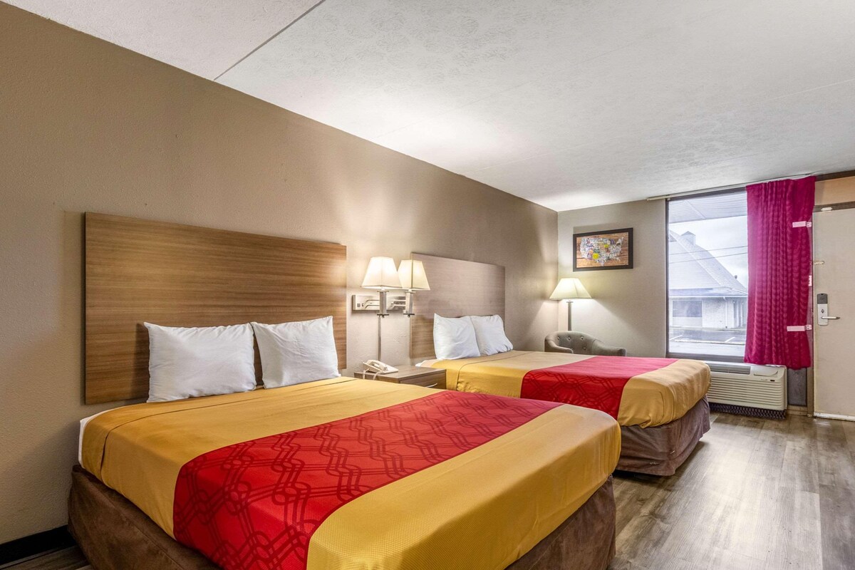 A hotel room with two beds, each with a headboard, bedside lamps, and a window with a curtain.
