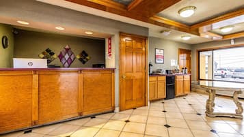 A hotel lobby with wooden reception desks, a glass-top table, and a view of the parking area through the door.