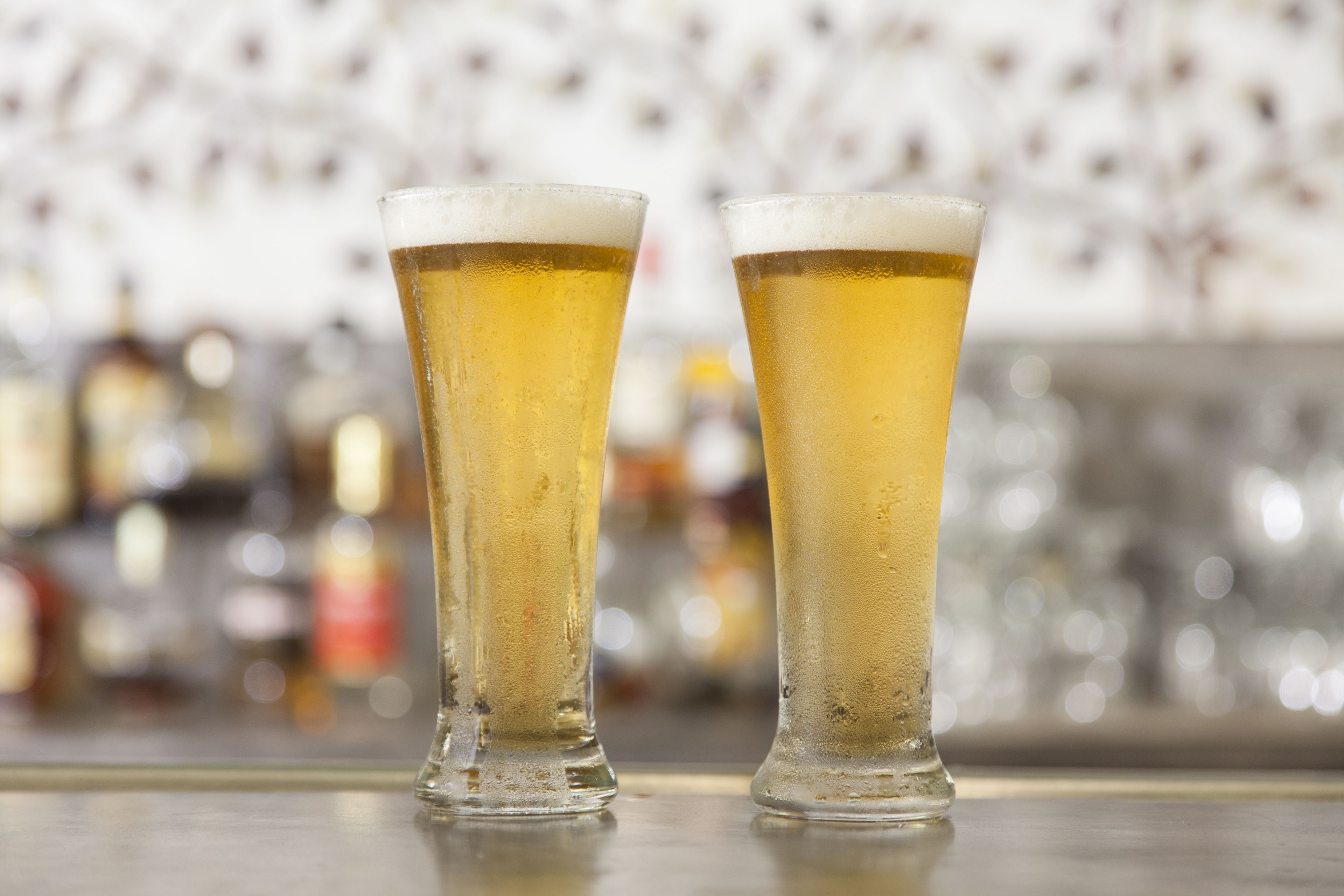 Two tall glasses of beer on a bar counter with a blurred background of bottles.