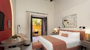 Ein Hotelzimmer mit einem großen Bett, einem Schreibtisch mit Stuhl, einem kleinen Tisch mit Pflanze und Blick ins Freie.