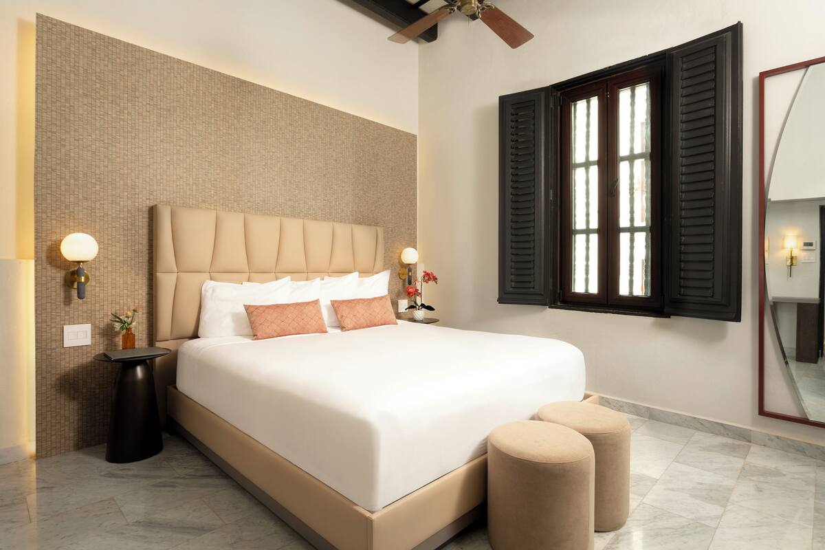 A modern hotel room with a large bed, a nightstand, a wall-mounted mirror, and a window with shutters.