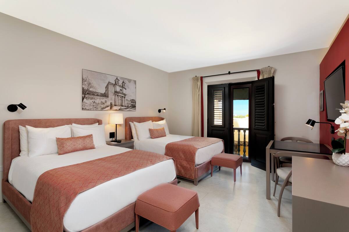 A hotel room with two beds, a desk, a chair, a lamp, and a painting on the wall.