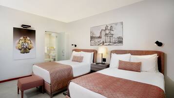 A hotel room with two beds, a bench, a bedside table with a lamp, and artwork on the walls.