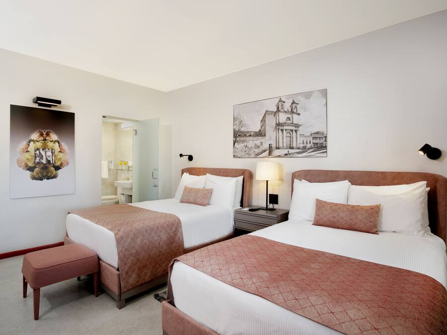 A hotel room with two beds, a bench, a bedside table with a lamp, and artwork on the walls.