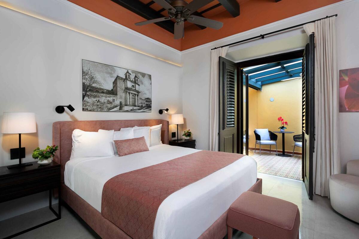 A hotel room with a large bed, two bedside tables with lamps, a ceiling fan, a painting on the wall, and an open door leading to a balcony with chairs and a table.