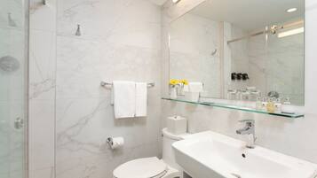 Une salle de bain moderne avec une douche à parois de verre, des toilettes blanches, un lavabo avec un miroir au-dessus et une étagère en verre où sont rangés les articles de toilette.