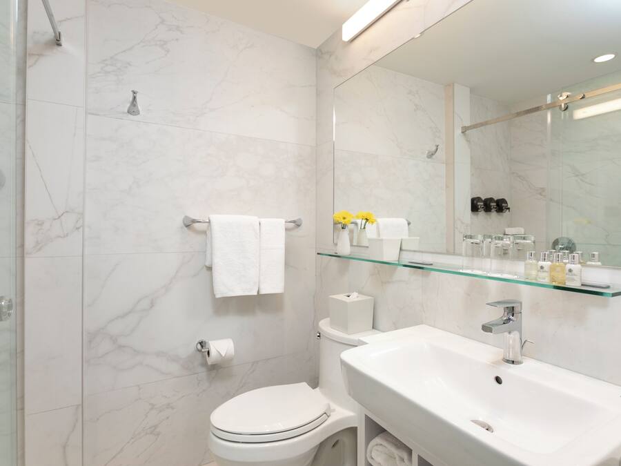 Une salle de bain moderne avec une douche à parois de verre, des toilettes blanches, un lavabo avec un miroir au-dessus et une étagère en verre où sont rangés les articles de toilette.