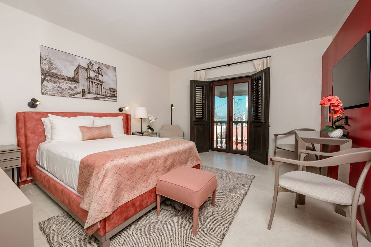 A bedroom with a bed, bedside tables, a chair, a desk, and a balcony with a view.