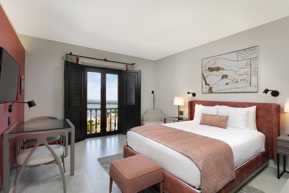 A hotel room with a bed, a desk, a chair, a nightstand, and a view of the outdoors.