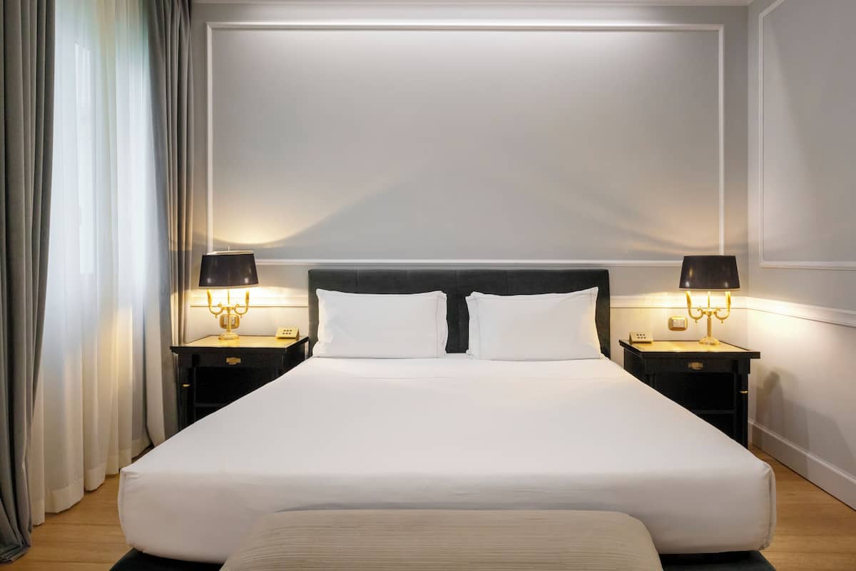 A neatly made bed with white linens, two bedside tables with lamps, and a large window with curtains.