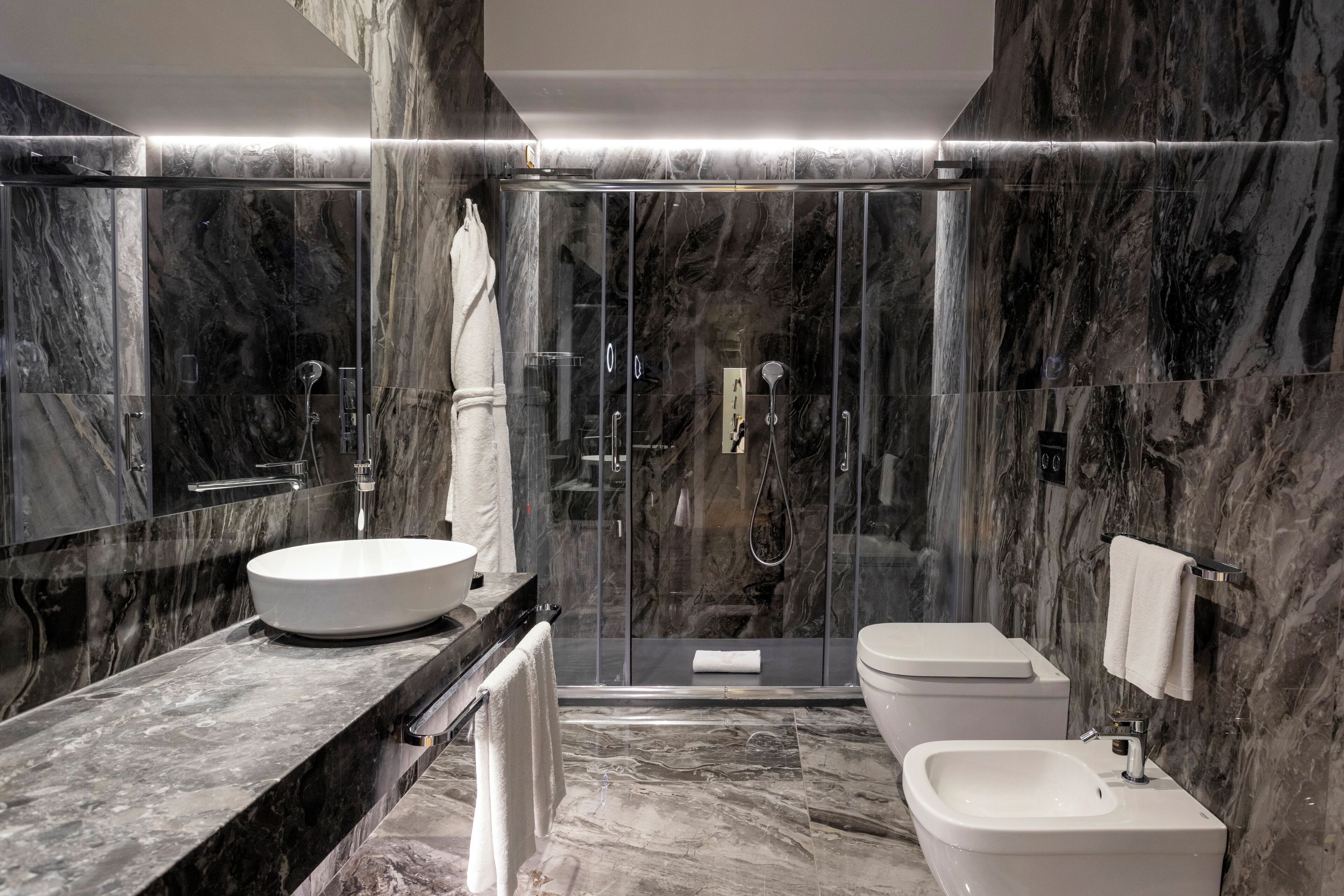 Une salle de bain moderne dotée d’un grand lavabo, de toilettes et d’un espace douche, avec des murs en marbre et une douche à parois de verre.