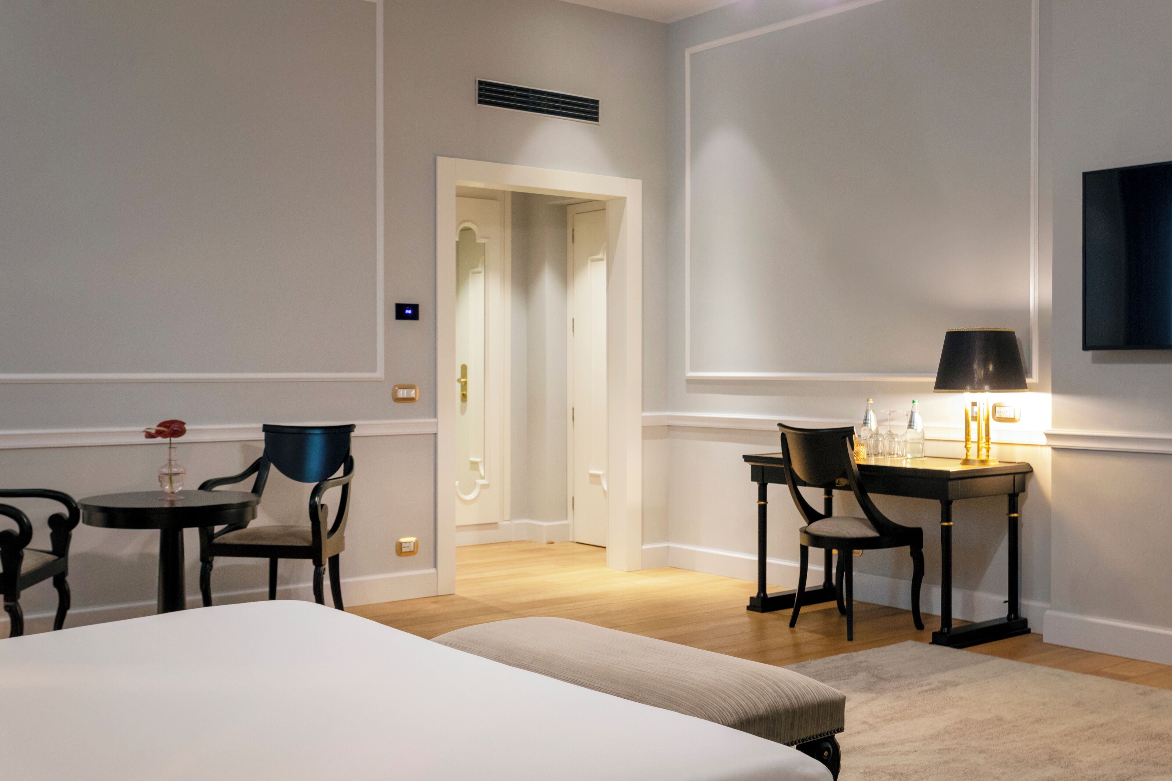 A hotel room with a bed, two chairs, a small table with a lamp, and a desk with a chair. There is a door leading to another room.