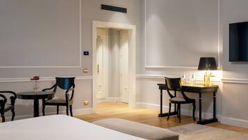 A hotel room with a bed, two chairs, a small table with a lamp, and a desk with a chair. There is a door leading to another room.
