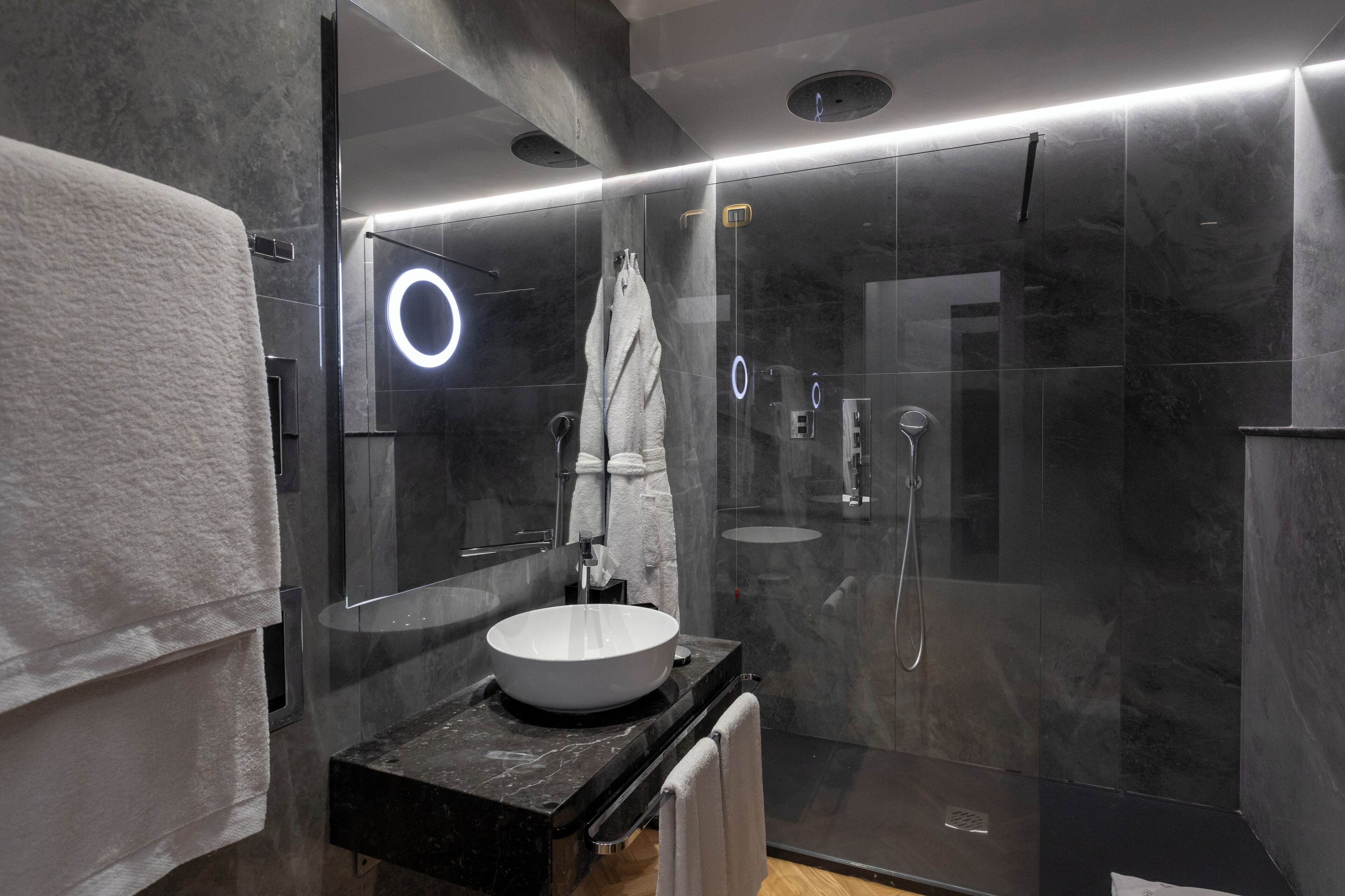A modern bathroom with a large mirror, a round light fixture, a glass-enclosed shower, and a black countertop with a white sink.