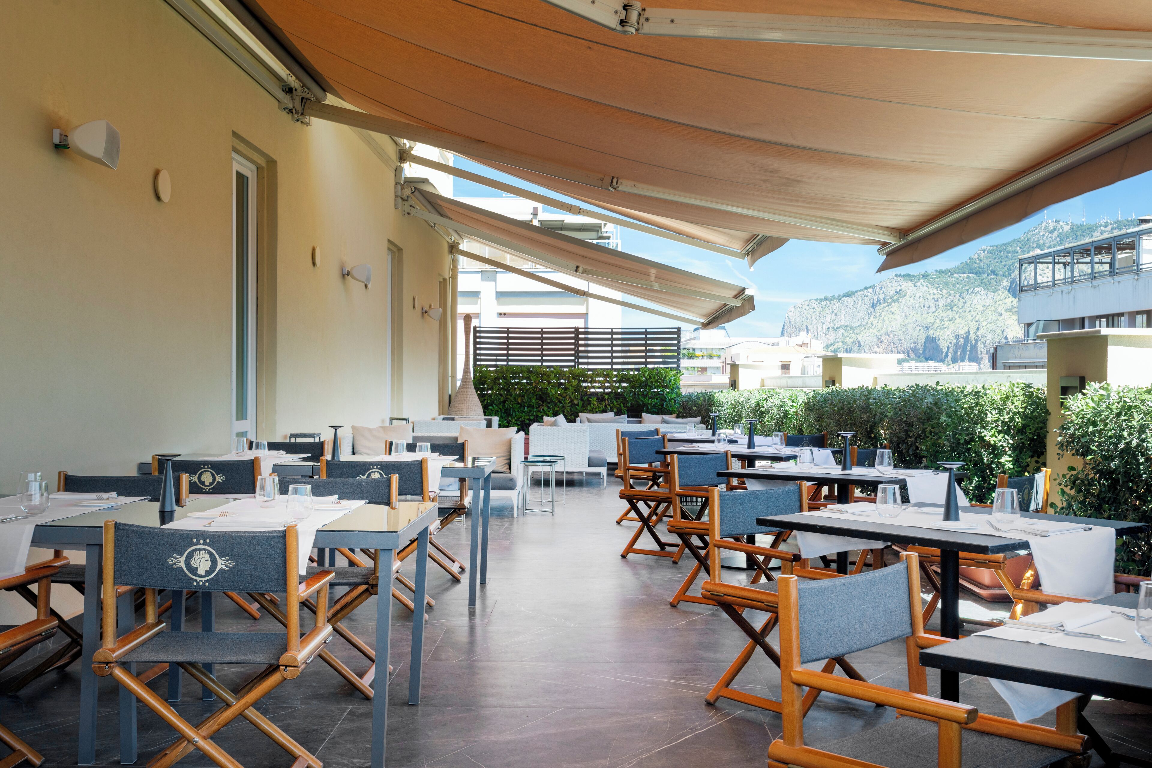 An outdoor dining area with tables and chairs, a covered walkway, and a view of buildings and greenery in the background.