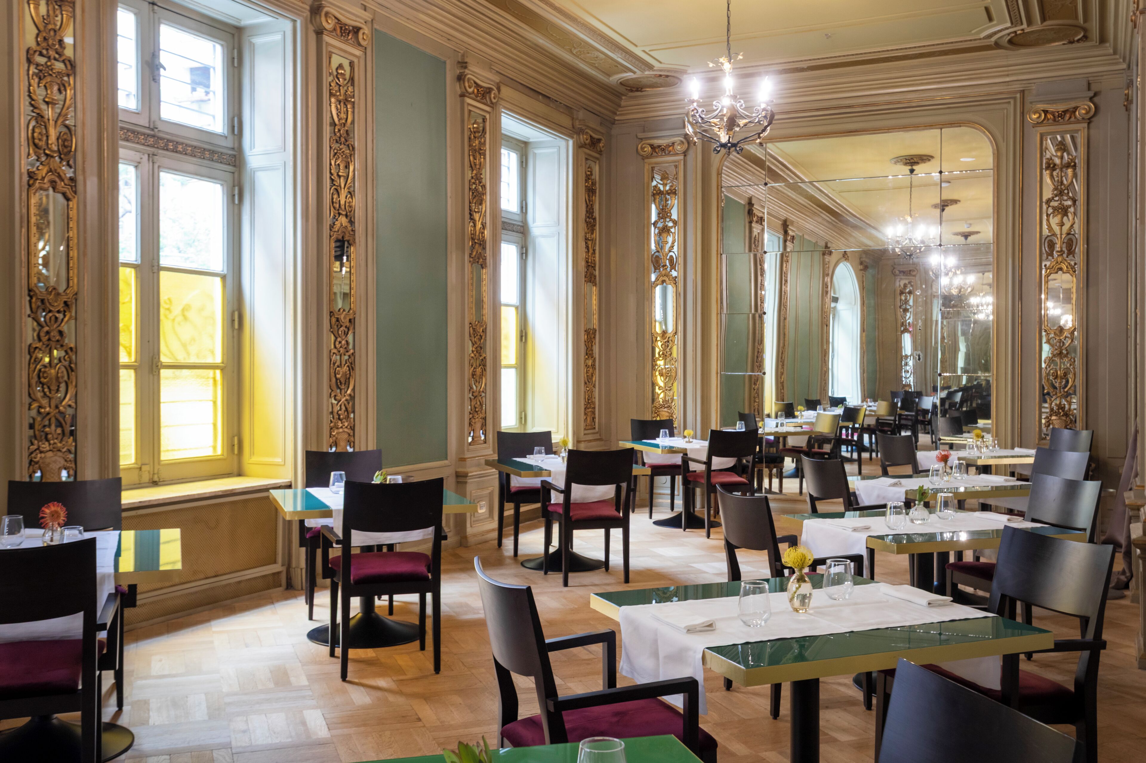 A spacious dining area with multiple tables and chairs, ornate wall decorations, and large windows allowing natural light.