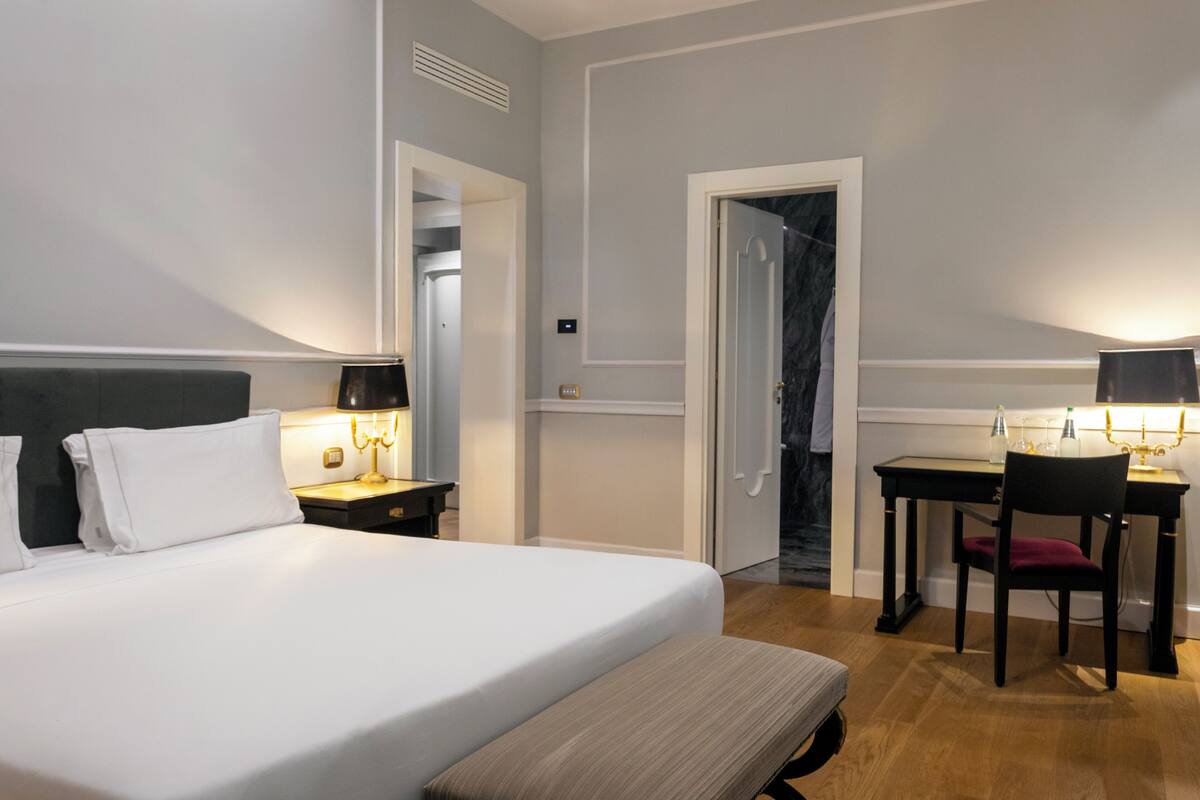 A hotel room with a bed, bedside table, lamp, desk, chair, and a door leading to a bathroom.