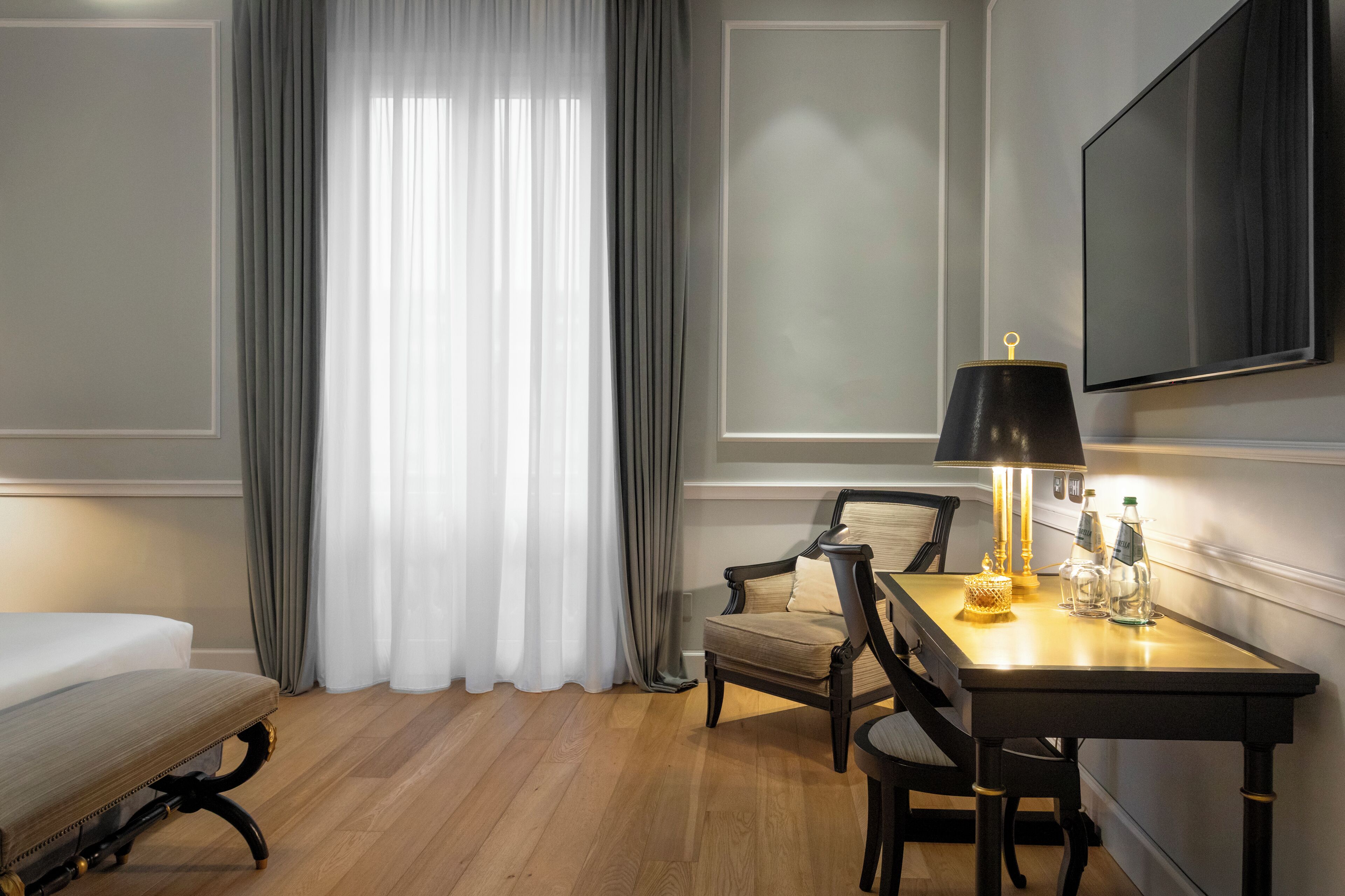 A hotel room with a bed, a desk with a lamp, a chair, and a TV mounted on the wall.