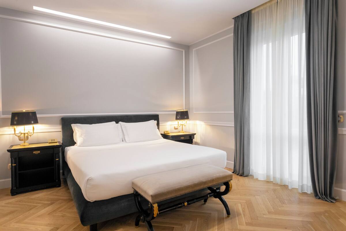 A neatly arranged hotel room with a large bed, two bedside tables with lamps, a bench at the foot of the bed, and a window with curtains.